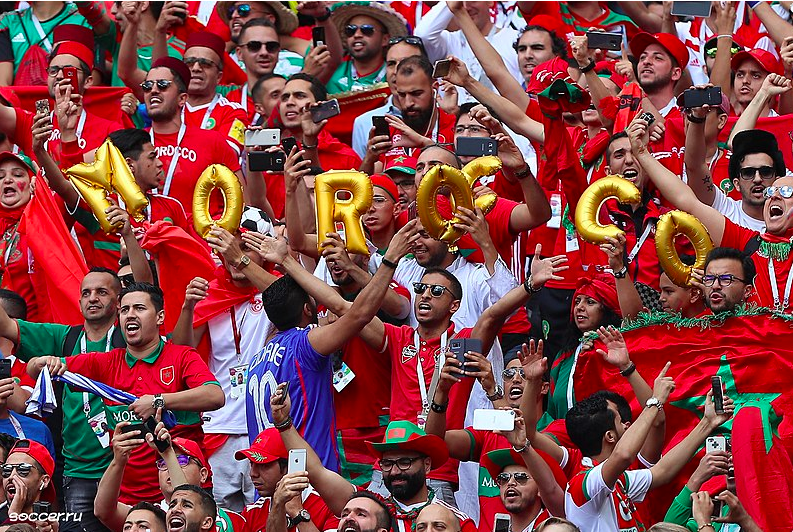A crowd of about 60 Moroccan football fans standing shoulder to shoulder in rows, in what appears to be stadium seating. All the fans are standing, and one row holds a set of gold, inflatable letters that read MOROCCO.